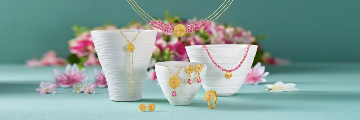 Goldschmuck mit rosa Edelsteinen, darunter Halsketten, Ohrringe und Ring, dekorativ arrangiert vor floralem Hintergrund als hochwertige Geschenkidee.